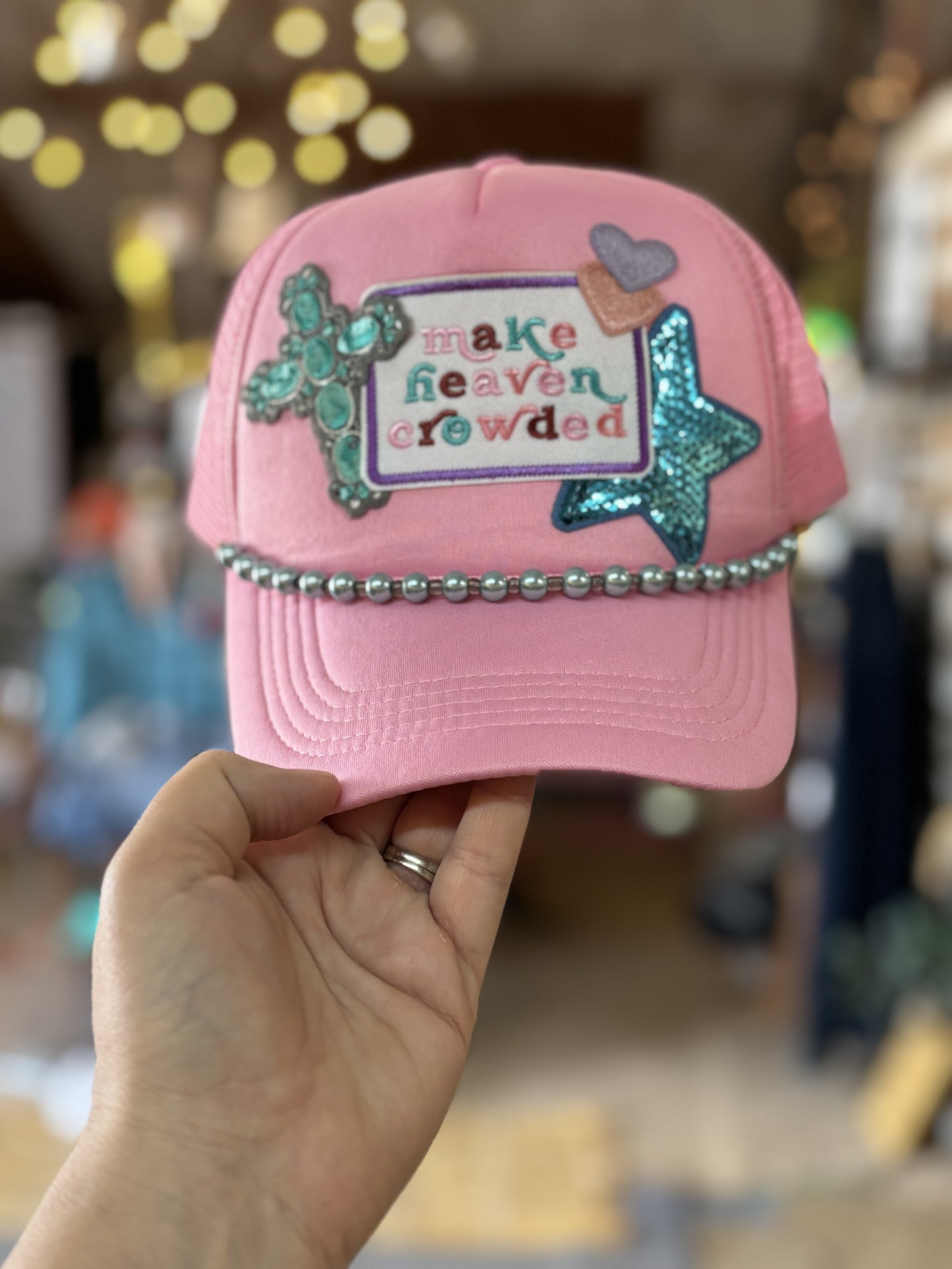 A pink trucker hat with the phrase 'make heaven crowded' written on it, adorned with blue and silver embellishments and a chain around the base.