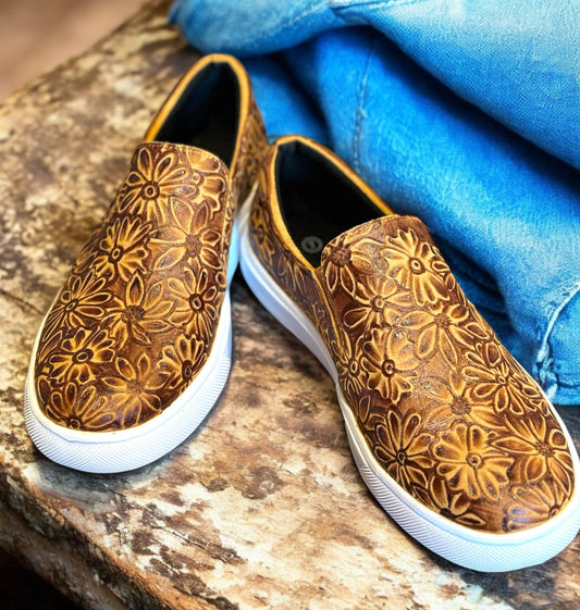 A pair of hand burned slip-on sneakers with a floral pattern on a brown background, displayed in front of a denim-covered object.